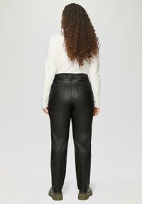 Schwarze Lederhosen mit hohem Bund und glatter Textur, zwei Gesäßtaschen und geraden Beinen, kombiniert mit einem weißen Langarmoberteil und grünen Stiefeln.