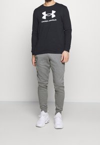 Schwarzes Langarmshirt mit weißem "Under Armour"-Logo, kombiniert mit hellgrauen, taillierten Jogginghosen und weißen Sportschuhen. Einfaches Design.