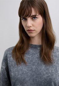 Młoda kobieta o prostych brązowych włosach z grzywką, ubrana w wyblakłą szarą bluzę, patrząca przed siebie z neutralnym wyrazem twarzy.