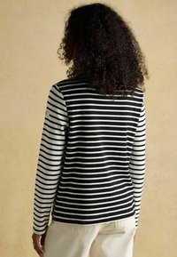 Pikem varrukaga topp, millel on mustad ja valged horisontaalsed triibud, valmistatud pehmest kangast. Vasakul varrukal on väike logopatch.