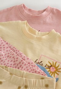 Deux sweats pour enfants pliés, un rose et un jaune avec un motif de soleil « hola sol », assortis à un pantalon rose à pois et un pantalon beige à motif solaire.