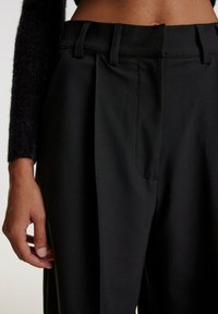 Schwarze, maßgeschneiderte Hose mit glatter Textur, Vorderfalten, Seitentaschen und hohem Bund. Auf dem Bild liegt eine Hand auf dem Stoff.