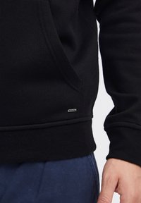 La sudadera negra presenta una tela texturizada, un bolsillo canguro y un pequeño acento de logotipo metálico cerca del dobladillo. Ajuste entallado con puños acanalados.