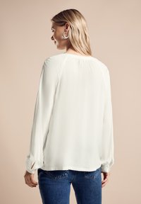 Witte blouse met lange mouwen, losse pasvorm, ronde halslijn en gedetailleerde plooien op de schouders. De stof heeft een gladde, zachte textuur.