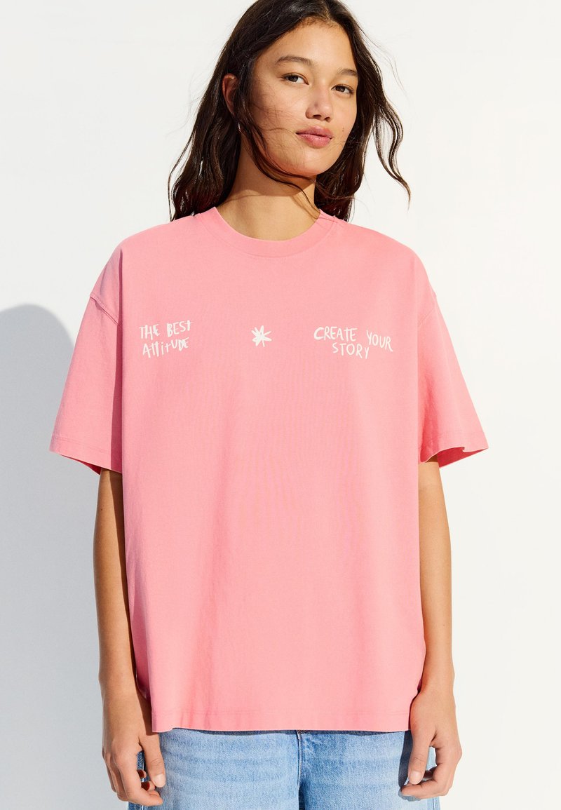 Jeune femme portant un T-shirt rose ample avec un texte blanc indiquant "La meilleure attitude" et "Créez votre histoire", assorti à un jean bleu clair.