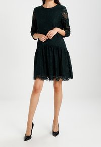 Dunkelgrünes Spitzenkleid mit dreiviertel Ärmeln, einem taillierten Oberteil und einem ausgestellten Rock. Es hat einen gewellten Saum und ein filigranes Spitzenmuster.