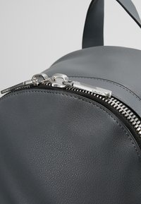 Sac à dos en cuir gris avec une forme arrondie, une texture lisse et deux fermetures éclair argentées avec le logo Calvin Klein en haut.