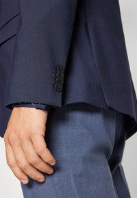 Navy blazer with a textured fabric, featuring a four-button cuff, paired with light blue checked pants. Hand positioned on the blazer.