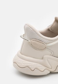 Beige sneaker med en texturerad stickad ovandel, vadderad krage och en gummisula med ett vågmönster; inkluderar varumärkesdetaljer och accenter.