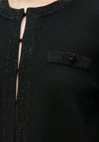 Μαύρο πλεκτό ένδυμα με λαιμόκοψη και τσέπη διακοσμημένες με χάντρες, καθώς και με ένα διακοσμητικό κουμπί. Η υφή φαίνεται απαλή και βελούδινη.