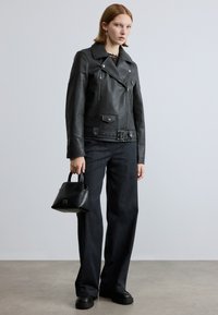 Schwarze Leder-Motorradjacke mit Reißverschlusstaschen, tailliertem Gürtel, kombiniert mit dunklen, weit geschnittenen Jeans und einer kleinen schwarzen Handtasche.