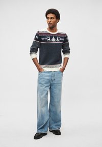 Námornícky sveter s ozdobnými vzormi s jeleňmi a vločkami; nosený s svetlomodrými džínsami širokých nohavíc a čiernou obuvou.