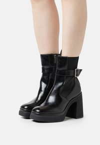 Schwarze Leder-Stiefeletten mit einem hohen Blockabsatz, seitlichem Reißverschluss und elastischem Einsatz. Sie besitzen einen Riemen mit einem metallischen Schnallen-Detail.