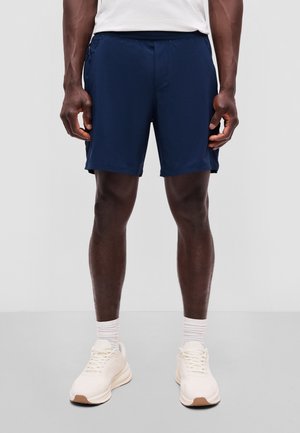 Mand iført marineblå træningsshorts, hvide sokker og off-white sneakers står foran en ensfarvet lys baggrund.