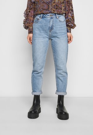 Person trägt hellblaue hochgekrempelte Jeans, schwarze klobige Stiefeletten und eine langärmelige gemusterte Bluse mit Rüschen an den Manschetten.