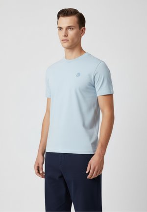 Giovane uomo con capelli corti castano chiaro, indossa una maglietta a maniche corte azzurro chiaro e pantaloni blu scuro, in piedi contro uno sfondo semplice.