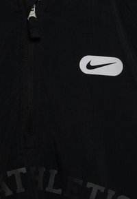 Chaqueta deportiva negra con cremallera, con un logo de Nike en blanco, que presenta un diseño de media cremallera y tela texturizada, acentuada por la palabra "ATHLETIC" en un estampado oscuro.