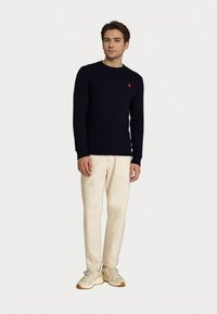 Young man standing wearing a navy cable-knit sweater, beige pants, and beige sneakers against a plain white background.