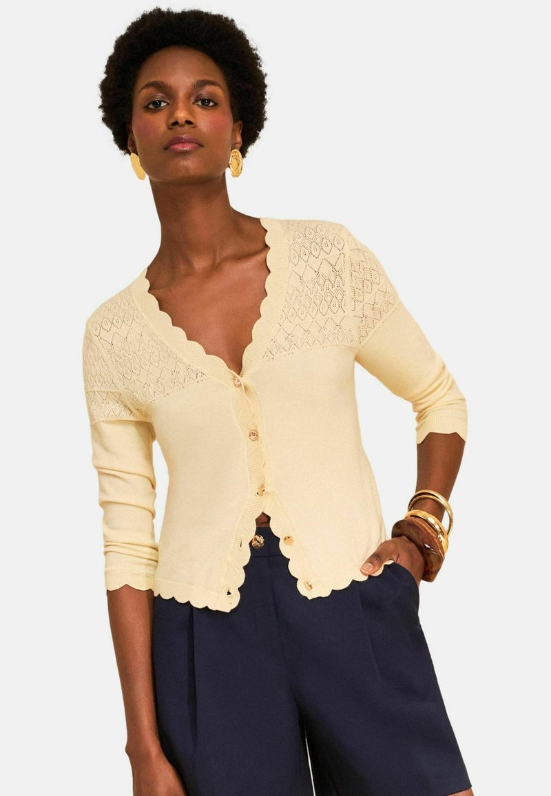 Nő krémszínű, gombos kardigánban, hullámos széllel és sötétkék, magas derekú rövidnadrágban, arany fülbevalókkal és karkötőkkel kiegészítve.