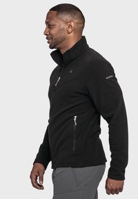 Schwarze Fleece-Jacke mit hohem Kragen, Seitenteilen und schlanker Passform. Mit einem Logo auf der Schulter, samtweicher Textur.