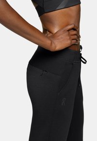 Pantalons de sport noirs avec une texture lisse, dotés de poches latérales zippées, d'une taille élastique avec cordon de serrage et d'un logo sur la cuisse.