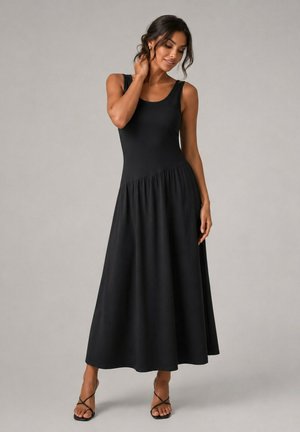 Femme posant dans une robe noire sans manches, ajustée, avec une taille asymétrique et une jupe évasée, portant des sandales à talons noires à brides.