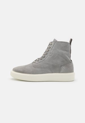 Basket montante en suède gris avec laçage à l’avant, semelle en caoutchouc blanche et languette arrière sur fond clair uni.