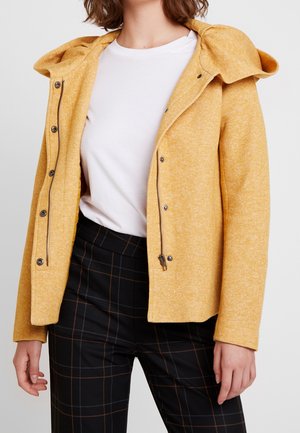 Kvinde iført en sennepsgul hættetrøje over en hvid t-shirt og sorte ternede bukser mod en ensfarvet baggrund.