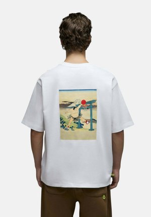 Personne aux cheveux bouclés portant un t-shirt blanc avec un sumo jouant au basket en plein air avec des montagnes et une porte torii en arrière-plan.