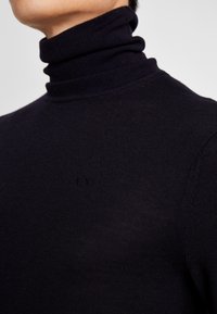 Sort rullekravesweater lavet af et let ribstrikket materiale med diskret logo broderet på brystet.