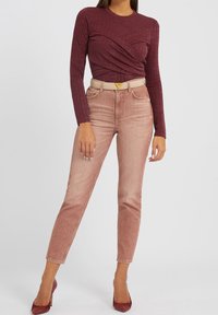 Bordeauxfarbenes Langarmoberteil mit strukturiertem Detail, kombiniert mit hellrosa Jeans und einem beigen Gürtel mit dreieckiger Schnalle; Modell trägt rote Absätze.