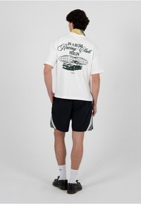 Homme portant un t-shirt blanc à motifs avec une voiture et le texte "In a Rush Racing Club Berlin", un short noir, des chaussettes blanches et des chaussures noires, debout de dos.