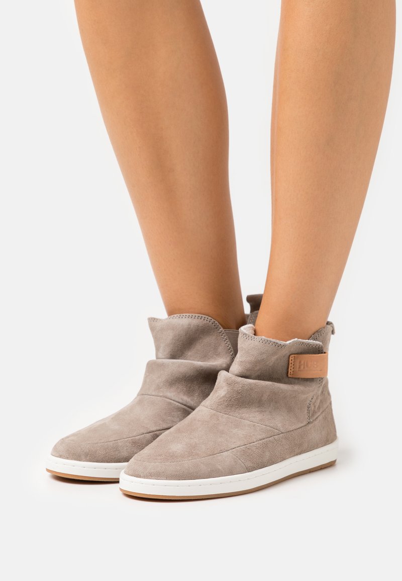 Cizme din suede gri până la gleznă, cu un design pliat, având un accent din piele bej și talpă din cauciuc alb. Textură netedă, cu un stil minimalist.