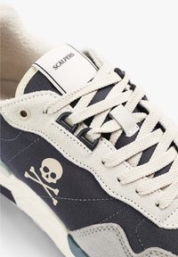 Tenisky s krémovým a námořnickým plátnem, semišovými akcenty, béžovými tkaničkami a logem lebky a zkřížených kostí na boku.
