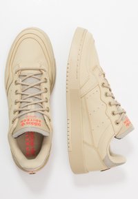 Béžové vysoké tenisky Adidas s síťovinovými prvky, polstrovaným límcem, silnou podrážkou a červeným značením "Adidas AdiTech" na jazyku a stélce.