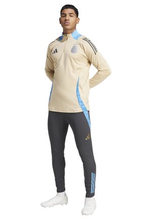 Beige sportjacket met blauwe accenten en rits, voorzien van een zwart logo. Grijze sportbroek met blauwe strepen en gouden branding.