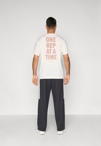 Világos bézs pamut póló, hátul rózsaszín felirattal: "EGY ISMÉTELÉS EGYSZERRE", fekete sportnadrággal, fehér oldalsávokkal.