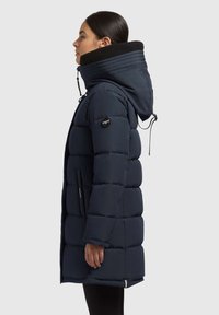 Mørkeblå puffjakke med høj krave, justerbar hætte og quiltet design. Har lynlåslommer på siden og et logo-patch på armen.