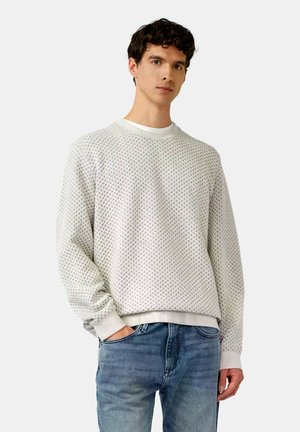 Jonge man met een lichtgrijze trui met patroon en blauwe spijkerbroek, staand met één hand in zijn zak tegen een effen achtergrond.