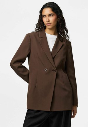 Femeie cu păr lung, creț și întunecat, purtând un sacou maro cu două rânduri de nasturi peste o cămașă albă și pantaloni negri, pozând cu o mână pe spate.