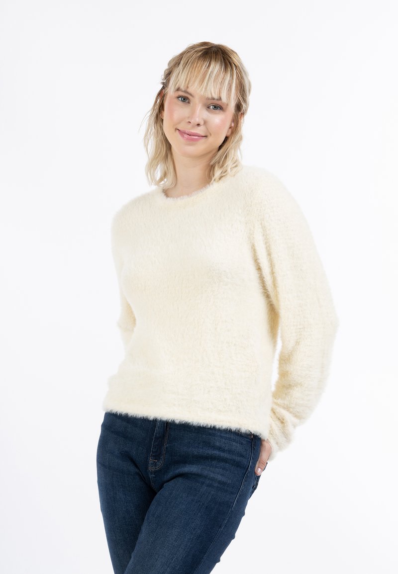 Maglione soffice di colore crema, con collo rotondo e maniche lunghe, abbinato a jeans blu scuro. Texture morbida, design casual.
