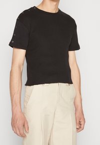Sort ribbet t-shirt med korte ærmer og cropped kant samt sølvøjede detaljer på skuldrene, stylet med lyse bukser.