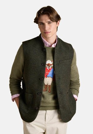 Junger Mann trägt einen olivgrünen Pullover mit einem Fuchs-Motiv, eine dunkelgrüne, geknöpfte Weste, ein rosa Hemd mit Kragen und hellbeige Hose.