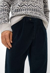 Pantaloni in corda blu scuro con chiusura a bottone, dotati di pence laterali e un tessuto testurizzato. Vestibilità affusolata, abbinati a un maglione lavorato a maglia con motivo.