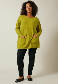 Grüner, gestrickter Pullover mit Rundhalsausschnitt, langen Ärmeln und zwei Fronttaschen, kombiniert mit schwarzen Skinny Jeans und schwarzen Flachschuhen.