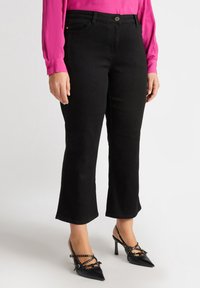 Schwarz ausgestellte Jeans mit Fronttaschen und einem Knopfverschluss, kombiniert mit schwarzen spitzen High Heels, die dekorative Riemen und Nieten aufweisen.