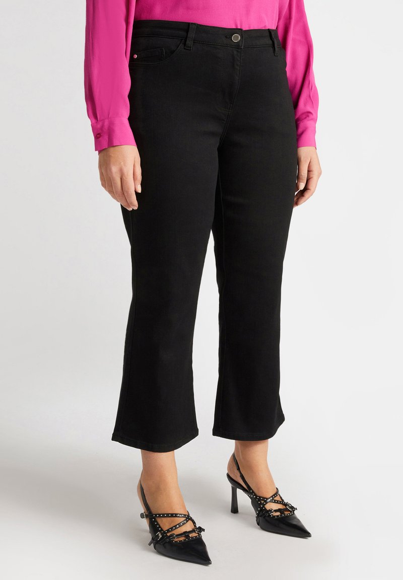 Schwarz ausgestellte Jeans mit Fronttaschen und einem Knopfverschluss, kombiniert mit schwarzen spitzen High Heels, die dekorative Riemen und Nieten aufweisen.