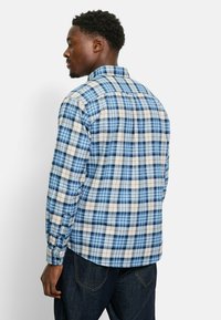 Blau kariertes Hemd mit Knöpfen, das eine Mischung aus hellblauen und dunkelblauen Quadraten mit weißen und orangen Akzenten zeigt. Lange Ärmel und ein Standardkragen.