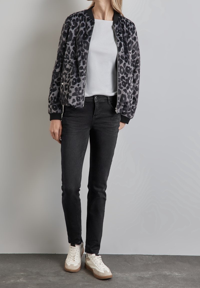 Femme debout portant une veste zippée à imprimé léopard, chemise blanche, jean noir et baskets blanches sur fond gris.