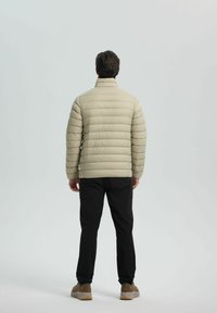 Beige pufferjack met hoge kraag, voorzien van horizontale stiksels, gedragen met zwarte broek en bruine sneakers, gezien van achter.